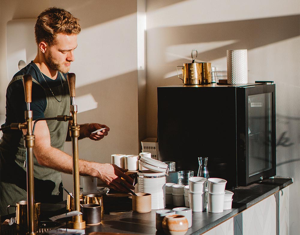 En barista som lager kaffe bak en disk mens sola skinner inn.